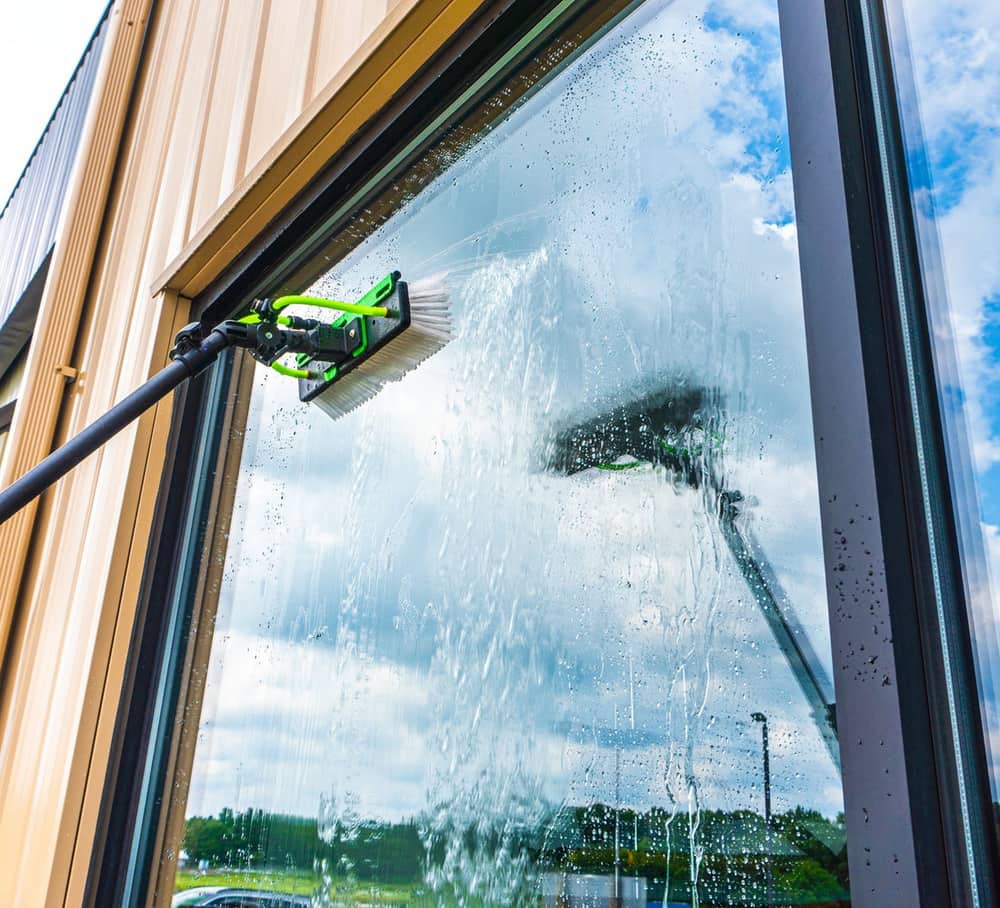 A water-fed pole with a soft brush head cleaning a tall window from the ground, pure water sheeting down the glass.